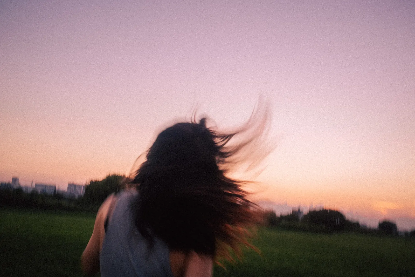 A blur of windswept dark hair against a violet dusk sky, a woman's shoulder just visible at the edge of the frame.