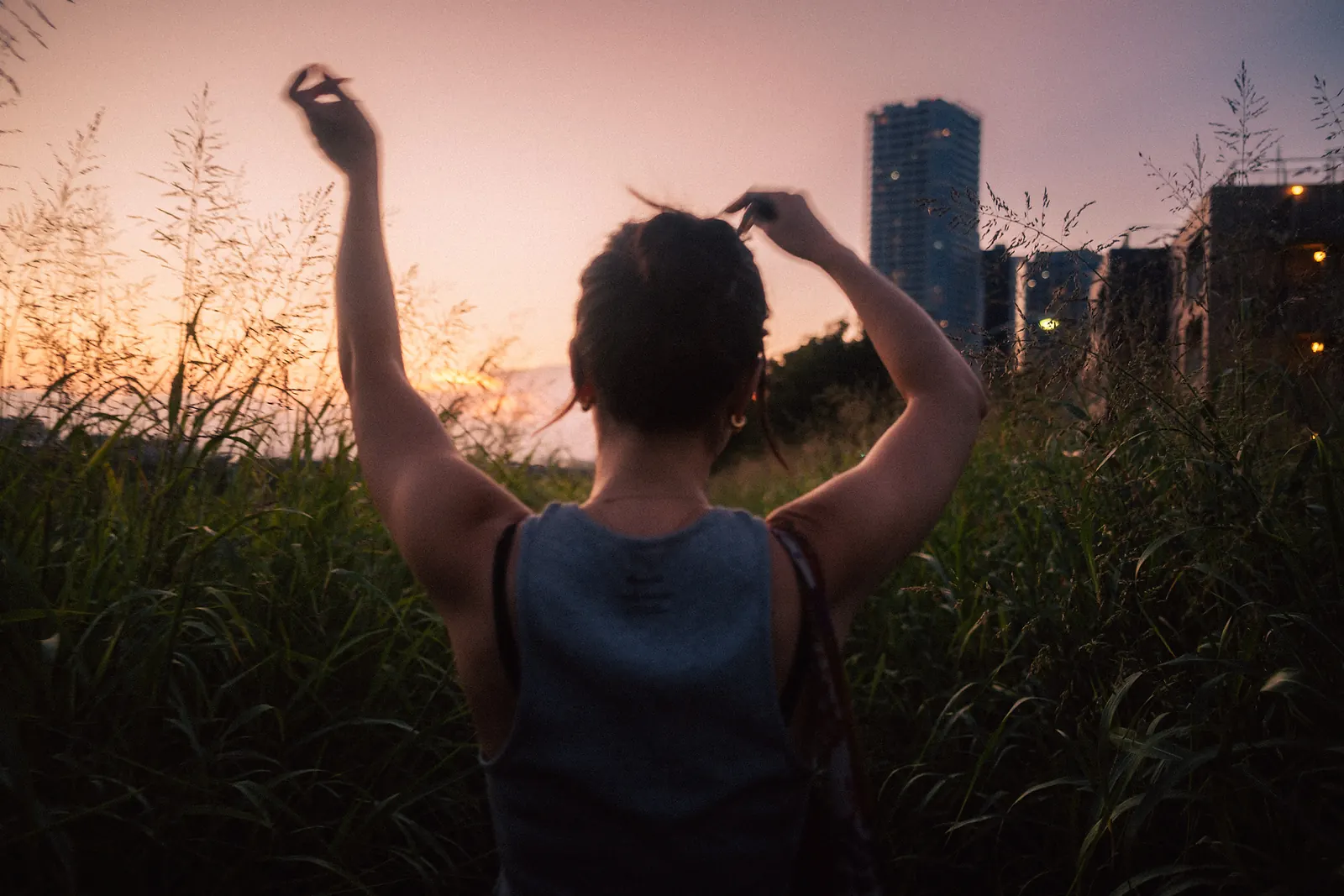 A woman seen from behind with both arms raised, gathering her hair, in tall grass against a pink dusk sky and apartment towers.