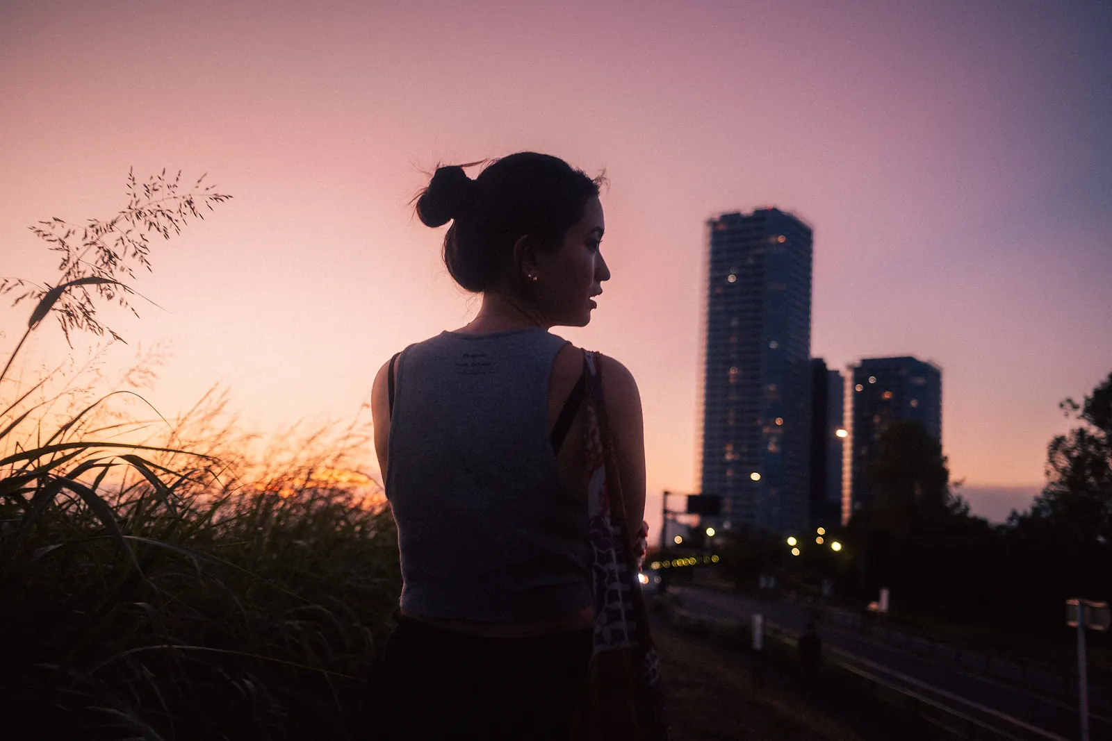 A woman in silhouette looking to the side, her hair in a bun, against a violet sunset sky with apartment towers and street lights.