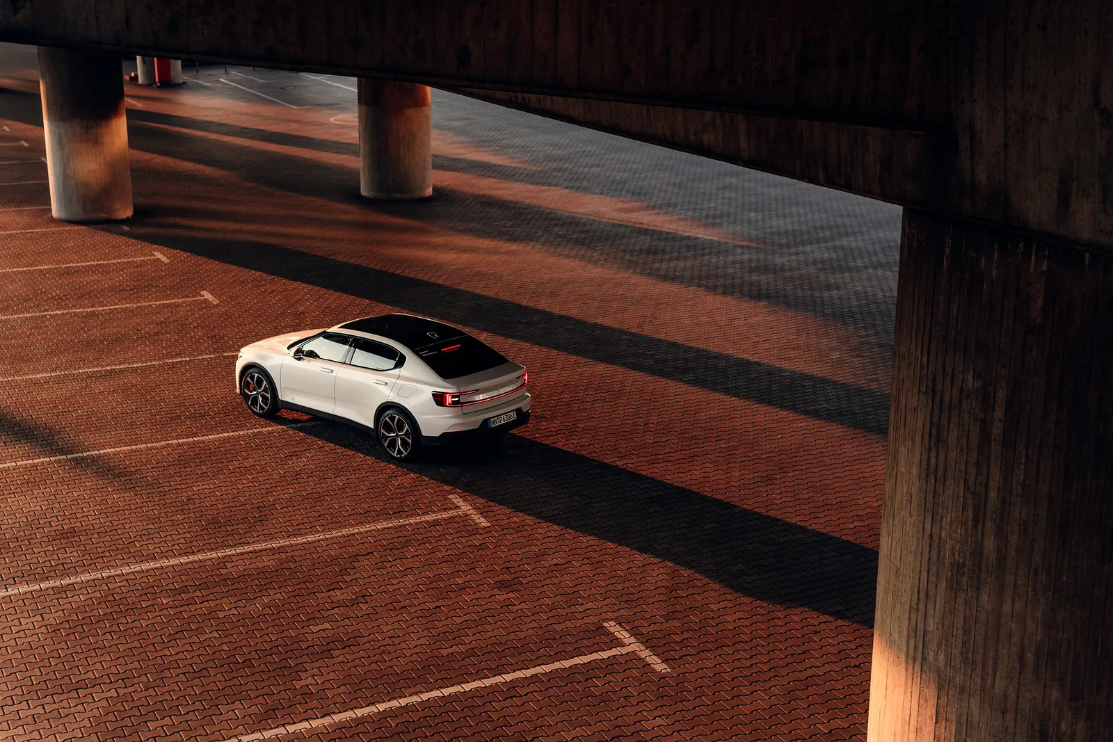 A white Polestar 2 parked in a brick-paved lot beneath a concrete structure, long shadows cast across the ground.