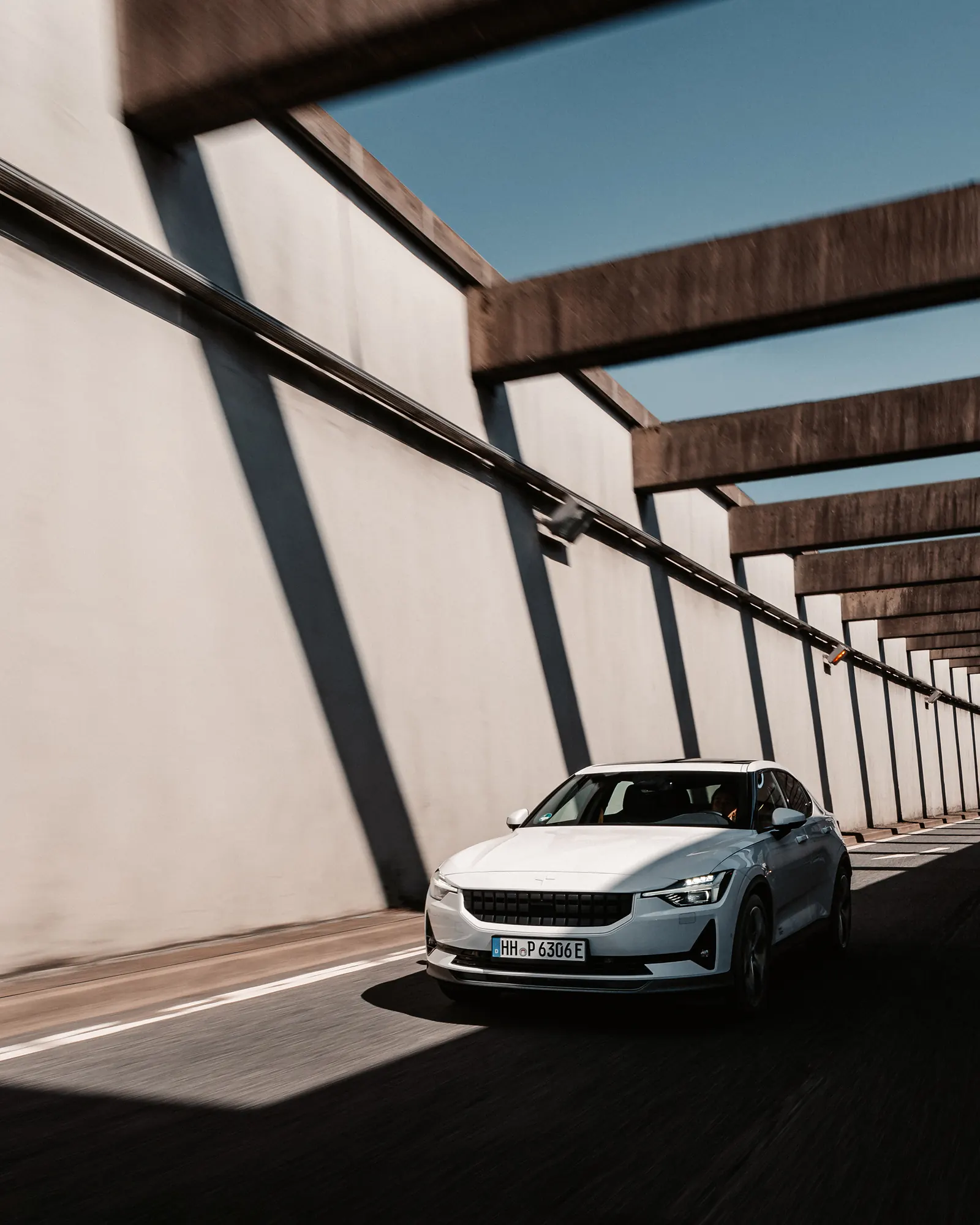 A white Polestar 2 driving along a parking-deck ramp beside a long concrete wall, beams casting diagonal shadows under a blue sky.