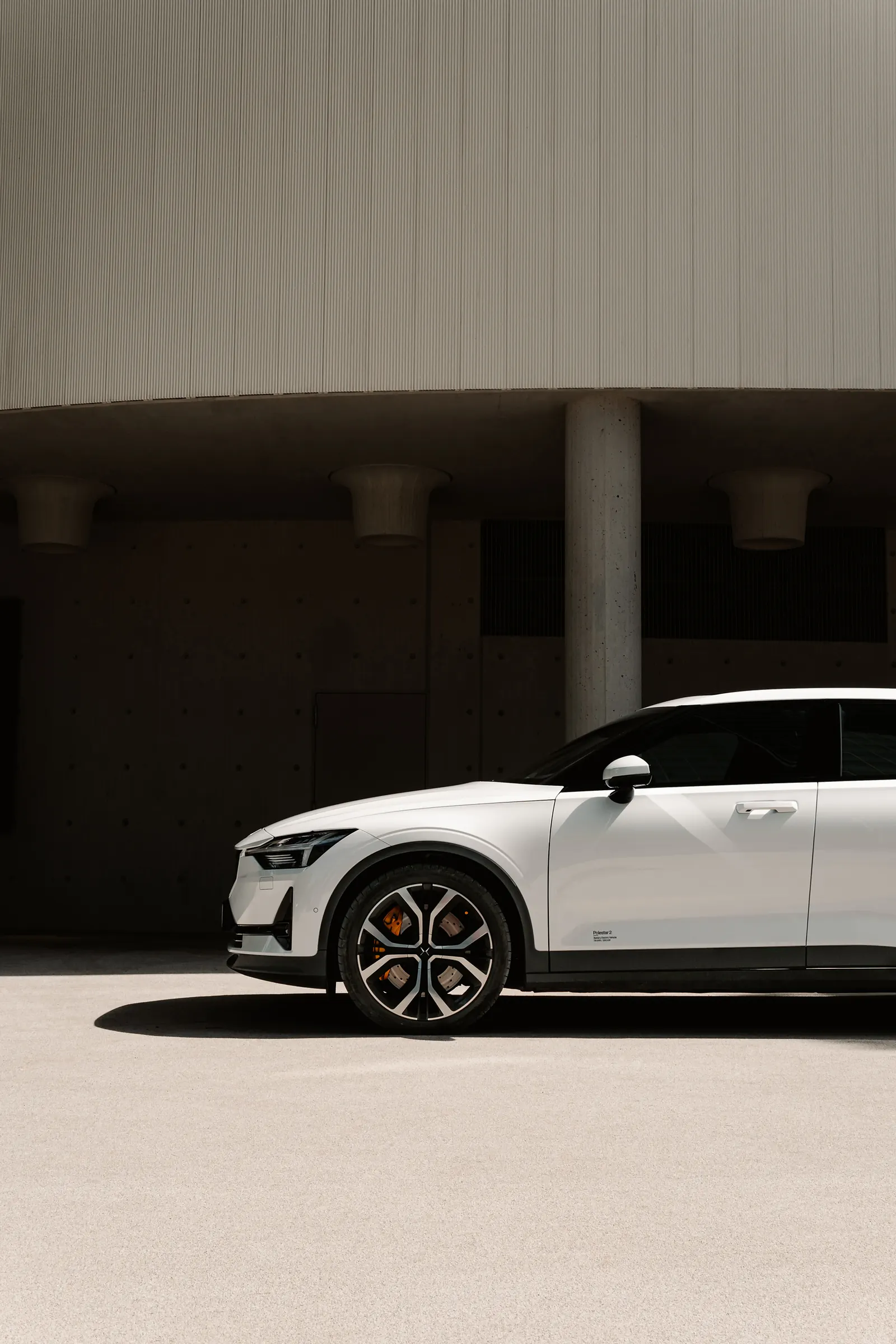 A white Polestar 2 in profile beneath the curved concrete overhang of a building, a single pillar behind it.