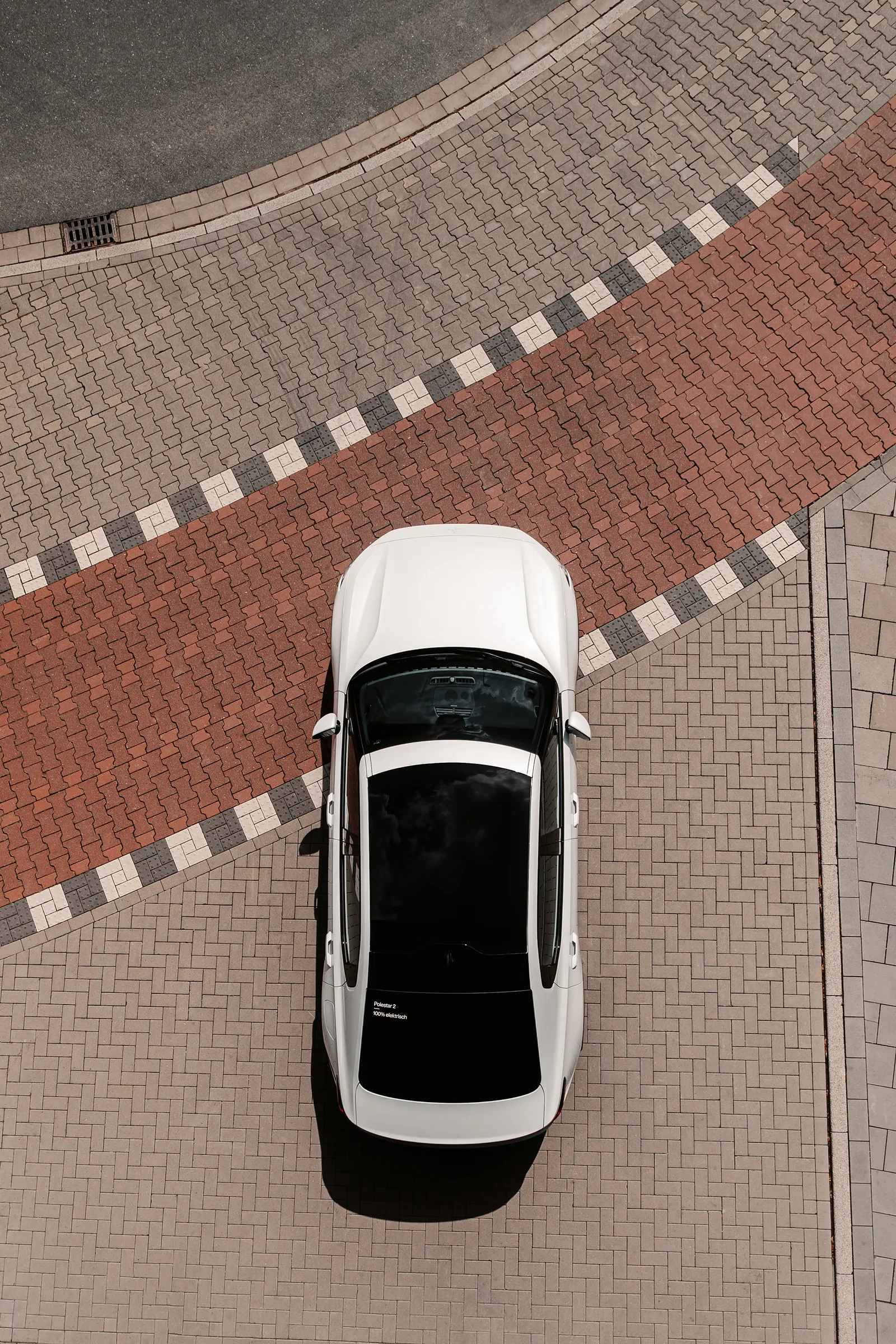 A drone shot looking straight down at a white Polestar 2 on a red-and-white striped brick plaza.