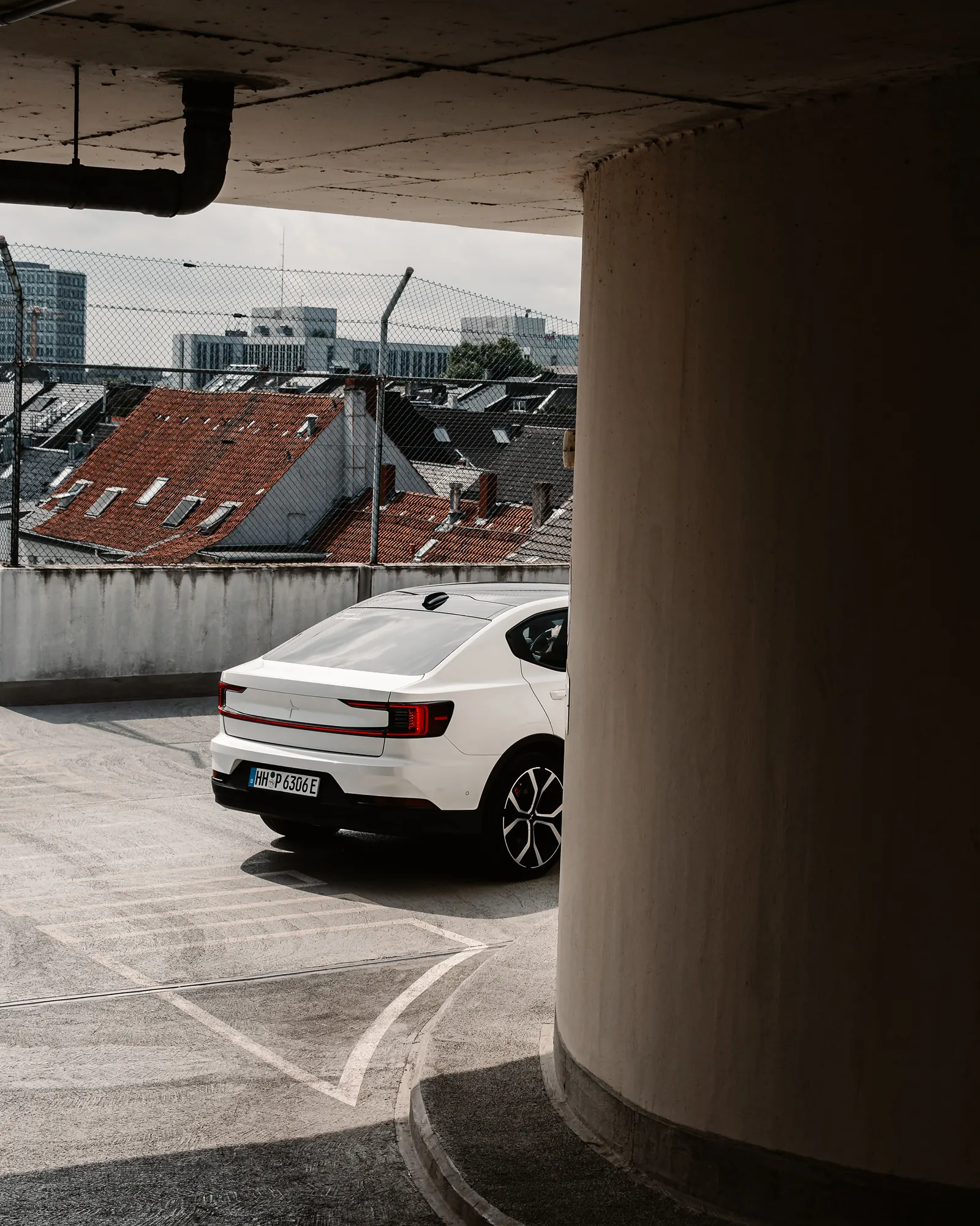 A white Polestar 2 seen from a rooftop parking deck beside a round concrete pillar, red-tiled old-town roofs visible beyond a chain-link fence.