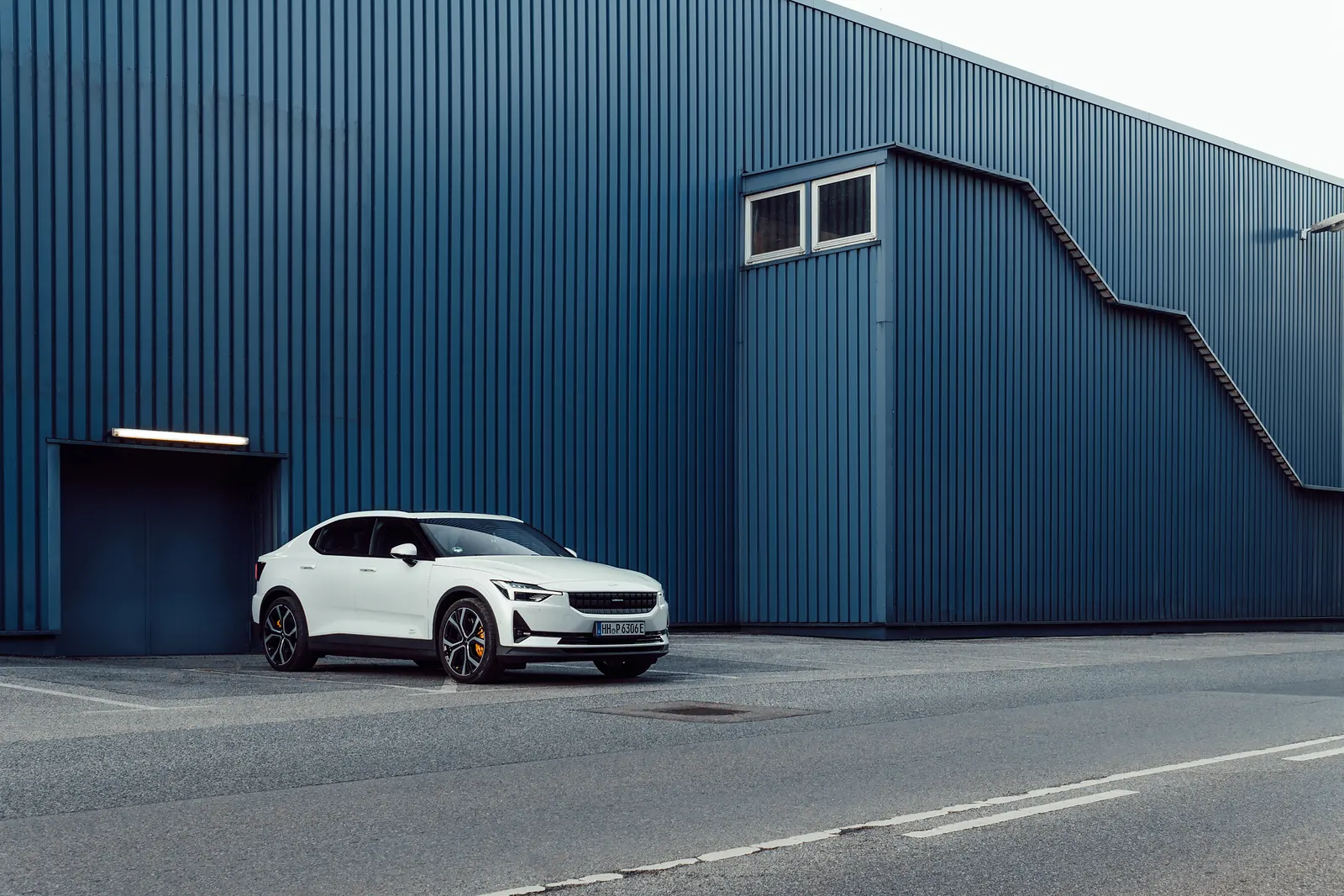 A white Polestar 2 parked in front of a warehouse with diagonal blue corrugated siding.