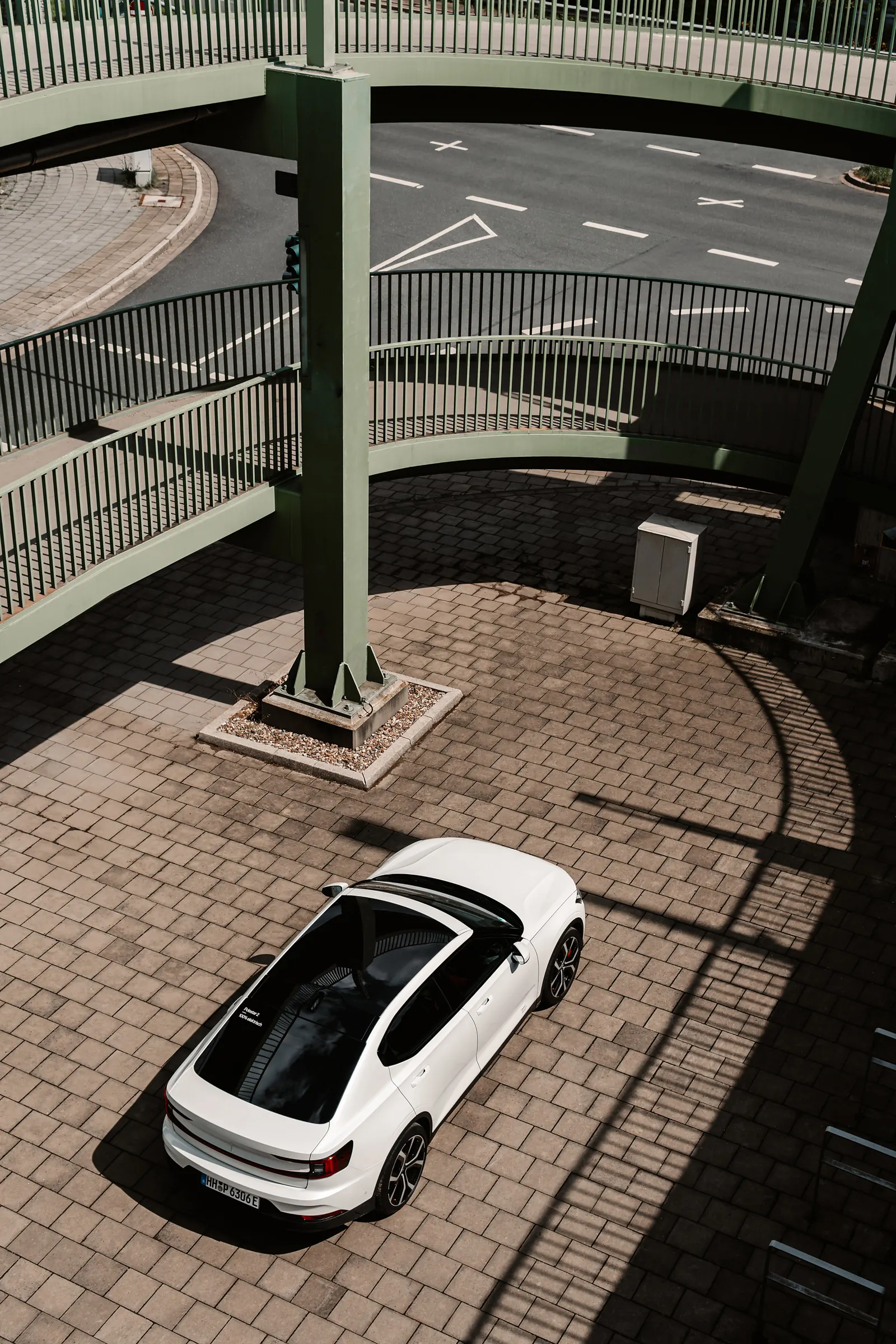 An aerial view down onto a white Polestar 2 parked beneath a curved pedestrian bridge.