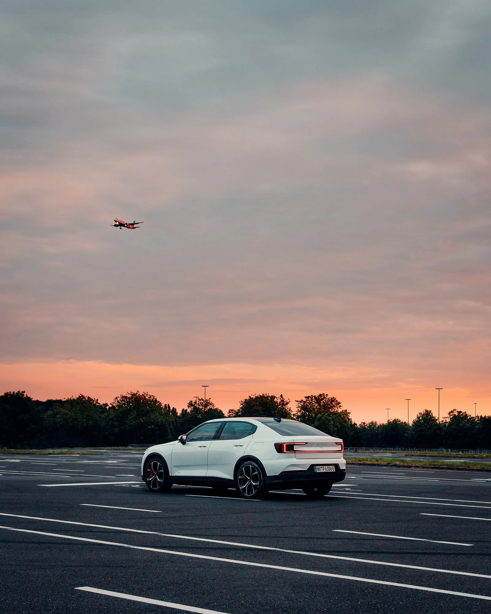 A white Polestar 2 parked on an empty lot at dusk, a plane crossing the pink-orange sky above.