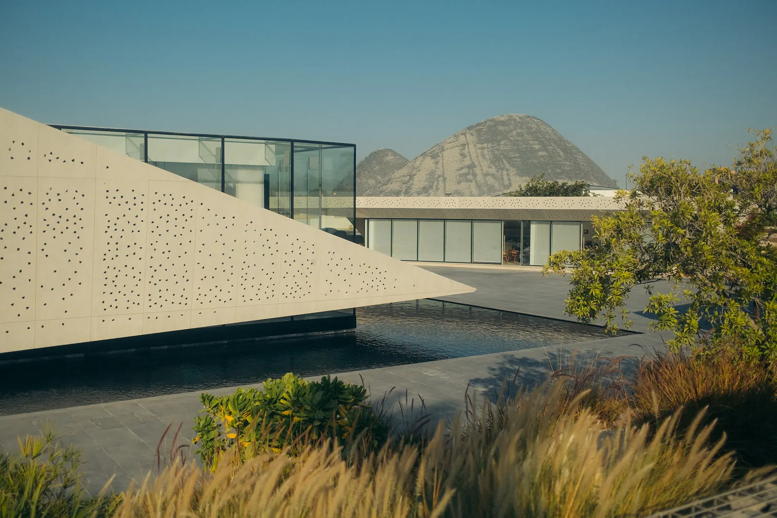 A perforated concrete pavilion beside a reflecting pool, with sculpted dune-shaped roofs in the distance.