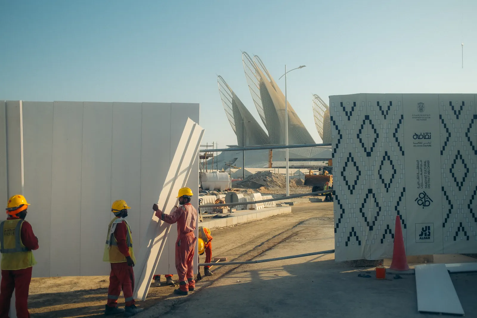 Construction workers carrying wall panels near a museum's steel falcon-wing towers, still under construction.