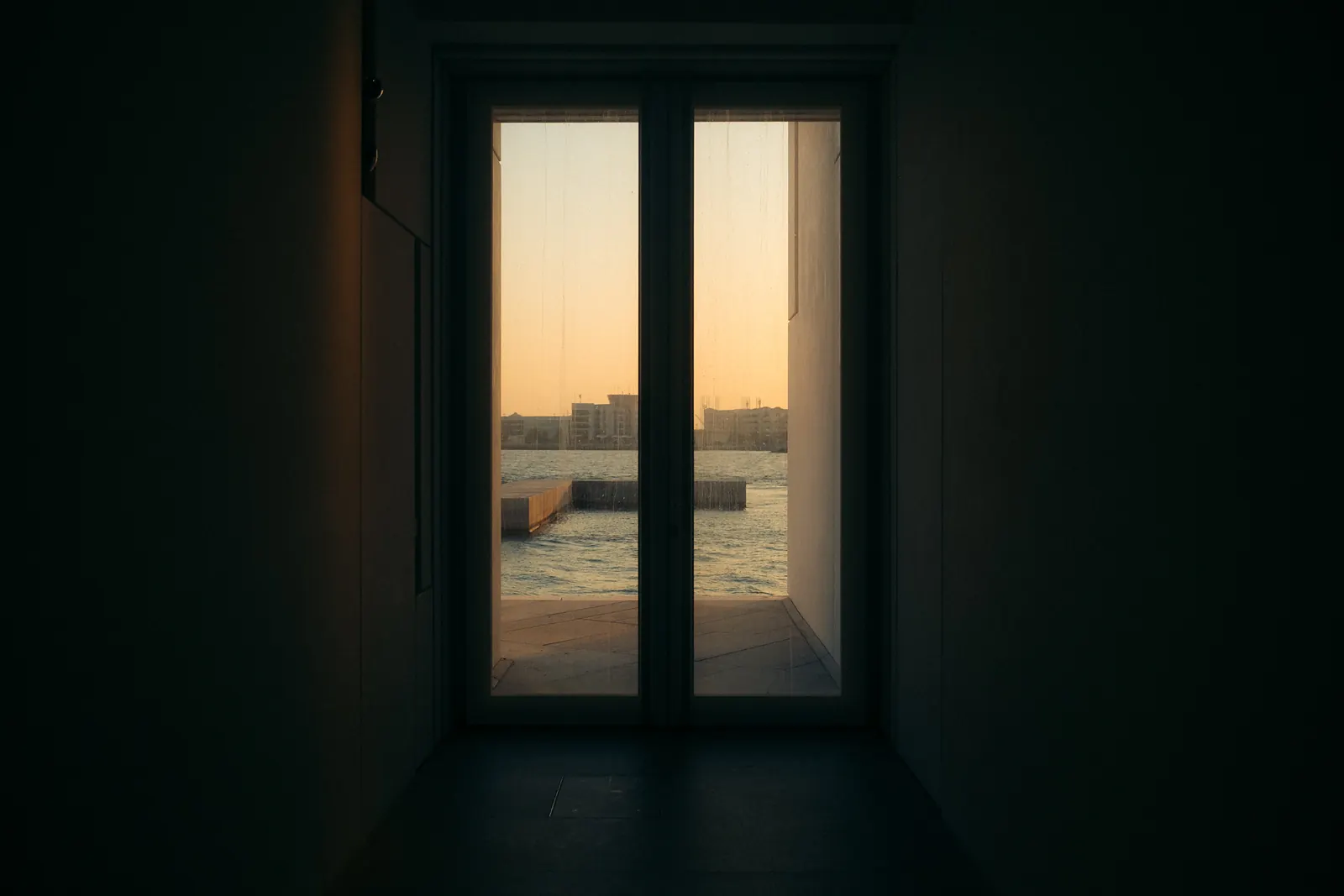 A dark corridor framing a glass door onto the waterfront, sunset light spilling in.