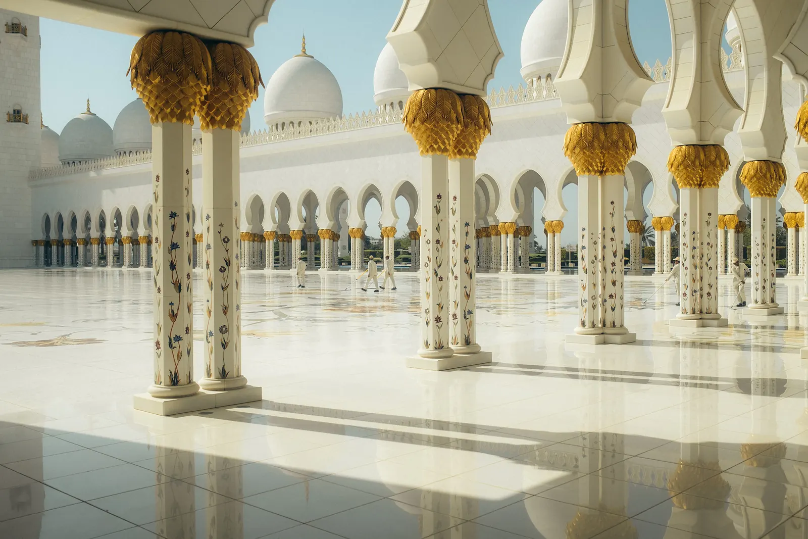 Gold-capped columns with floral inlay in the Grand Mosque's courtyard, staff cleaning the marble floor.
