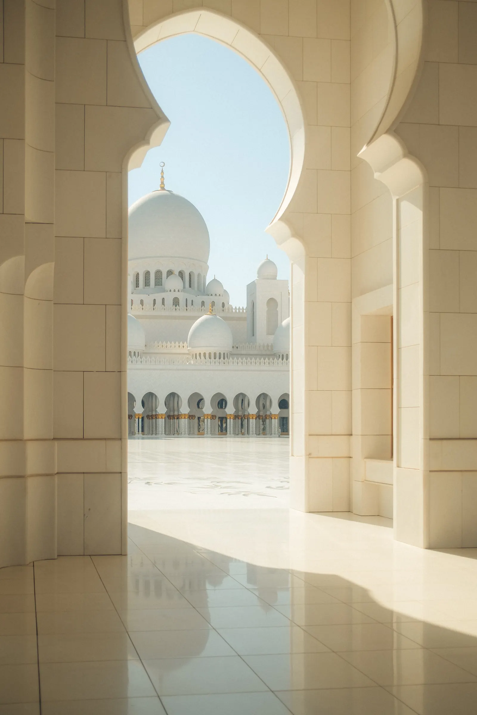 The white domes and colonnade of the Sheikh Zayed Grand Mosque seen through a keyhole arch, the courtyard reflected faintly on the polished floor.